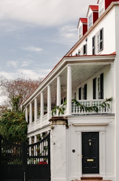 A large, white colonial-style house with a red roof and multiple chimneys is shown. The house features a prominent front porch supported by white columns and decorated with green garlands. There are several tall windows with black shutters and a black door with an address number on it. Surrounding the house, there is greenery and a black iron gate at the entrance.