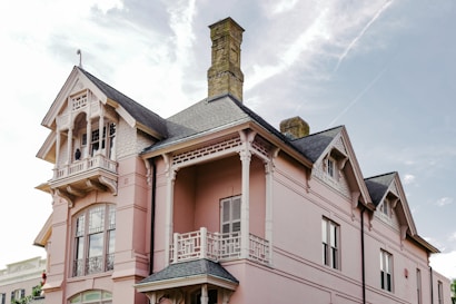 A large, elegant Victorian-style house painted in light pink with intricate architectural details. The building features ornate woodwork, gabled roof sections, and a prominent brick chimney. Large windows and a balcony add to the grandeur of the house, and the sky in the background is partially cloudy with a bright atmosphere.