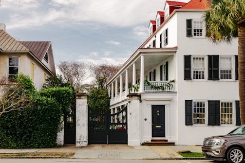 A large, white, two-story house with black shutters and a red roof is flanked by a smaller house with a sloped brown roof. Tall trees and a well-trimmed hedge are seen surrounding the property. An ornate white gate is prominently featured in front of the house. The setting appears to be calm and residential.