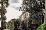 A wrought iron fence decorated with festive wreaths lines a sidewalk bordered by lush greenery and palm trees. Behind the fence, a row of classic and elegant townhouses with beige brick facades and large windows reflects a serene and upscale neighborhood. The bright sky overhead adds a sense of openness.