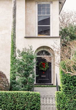 A beige stucco house facade with a tall, arched doorway. A dark front door is adorned with a green wreath featuring a red ribbon, set at the top of a short flight of steps with iron railings. Lush greenery, including trimmed hedges and trees, surrounds the entrance.