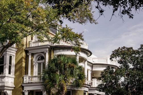 A large, elegant house with classic architectural details, such as white columns and intricate moldings, is surrounded by lush greenery. Tall trees with green leaves partially obscure the view of the building, adding to the serene atmosphere.