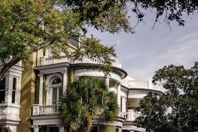 A large, elegant house with classic architectural details, such as white columns and intricate moldings, is surrounded by lush greenery. Tall trees with green leaves partially obscure the view of the building, adding to the serene atmosphere.