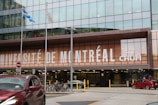 A modern building facade with large glass windows, displaying the words 'Université de Montréal CHUM' prominently in bold white letters. In front of the building, there are several bicycles parked and a red car passing by on the street. Two flags are flying on poles, and there is a no entry sign visible.