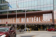 A modern building facade with large glass windows, displaying the words 'Université de Montréal CHUM' prominently in bold white letters. In front of the building, there are several bicycles parked and a red car passing by on the street. Two flags are flying on poles, and there is a no entry sign visible.