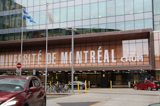 A modern building facade with large glass windows, displaying the words 'Université de Montréal CHUM' prominently in bold white letters. In front of the building, there are several bicycles parked and a red car passing by on the street. Two flags are flying on poles, and there is a no entry sign visible.