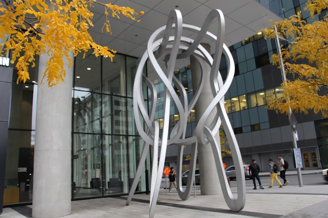 A modern abstract sculpture made of intertwining metallic loops stands outside a contemporary glass and concrete building. Autumn leaves in bright yellow frame the top of the image, providing contrast against the structural forms. Several people walk along the sidewalk nearby, dressed in casual attire.