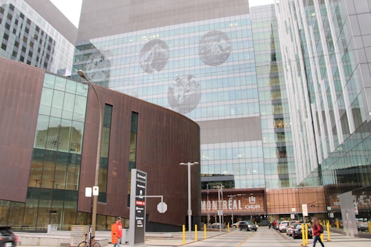 A modern architectural structure featuring large reflective glass windows and metal panels. The facade includes artistic circular images that resemble celestial bodies. The building displays the word 'Montreal' and 'CHUM' in bold letters. Pedestrians are visible in the foreground, along with parked cars and street signs.