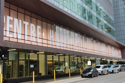 A modern building facade with a large glass front, displaying the text 'CENTRE HOSP...' partially visible. Several cars are parked in front of the building on a paved area with yellow bollards. The architecture features glass and metal elements with reflections visible on the windows.