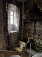 A cozy, eclectic interior with exposed brick walls and a vintage vibe. A narrow window allows natural light to filter in, highlighting ornate patterned curtains. There's a comfortable, colorful sofa adorned with various patterned cushions. A matching lamp with a decorative shade stands beside the sofa. The patterned wallpaper adds to the warm, artistic atmosphere.