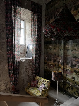 A cozy, eclectic interior with exposed brick walls and a vintage vibe. A narrow window allows natural light to filter in, highlighting ornate patterned curtains. There's a comfortable, colorful sofa adorned with various patterned cushions. A matching lamp with a decorative shade stands beside the sofa. The patterned wallpaper adds to the warm, artistic atmosphere.