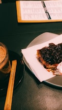 A close-up view of a dessert with a dark, glossy topping placed on a white paper atop a gray plate. Beside it, there's a cup of coffee on a saucer, along with a wooden stirrer. A menu is partially visible in the background on a black surface.