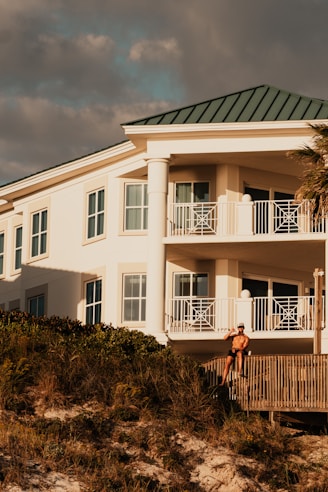 A luxury white villa with large windows and a green roof is situated on a scenic hillside. A man in sunglasses and swimwear is casually seated on a wooden railing, enjoying the warm sun.
