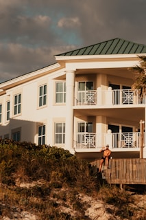 A luxury white villa with large windows and a green roof is situated on a scenic hillside. A man in sunglasses and swimwear is casually seated on a wooden railing, enjoying the warm sun.