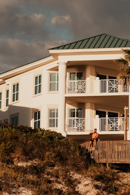 A luxury white villa with large windows and a green roof is situated on a scenic hillside. A man in sunglasses and swimwear is casually seated on a wooden railing, enjoying the warm sun.
