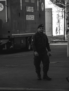 A person in military attire walking on a street near a large vehicle with German text on the side. The environment appears urban with other structures and people partially visible in the background.