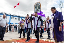 A group of men dressed in purple and white attire are participating in a religious procession, carrying a decorated religious icon adorned with purple and white fabrics and balloons. One woman in the background is holding a bouquet of flowers. There are several people in the background observing the event. The background includes a building with a blue and white facade and multiple windows, and a flagpole with a partially visible red and white flag.