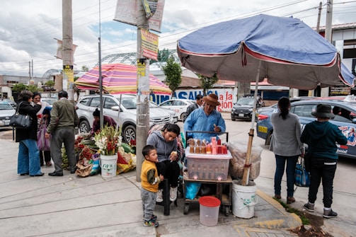 A busy street market scene with people gathered around a food vendor under a large blue umbrella. The vendor is serving drinks from a cart filled with colorful bottles. Nearby, a group of people are interacting next to flower vendors selling bouquets in buckets. Cars are passing by in the background, and various signs and posters are visible on the poles.