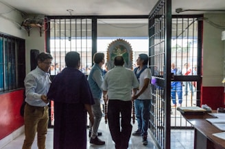 Several people stand near a large open gate in a building. Beyond the gate, a group of people can be seen outside, with a religious icon or statue partially visible. The setting appears to be a mix of indoor and outdoor environments, possibly a religious or community event. The interior has walls painted red and white, and there are desks and papers visible to the side.