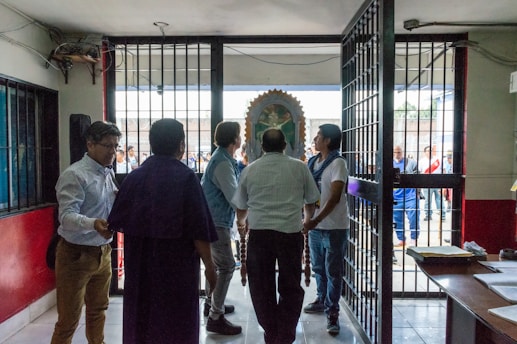 Several people stand near a large open gate in a building. Beyond the gate, a group of people can be seen outside, with a religious icon or statue partially visible. The setting appears to be a mix of indoor and outdoor environments, possibly a religious or community event. The interior has walls painted red and white, and there are desks and papers visible to the side.