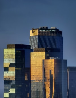 Several modern skyscrapers with reflective glass surfaces appear in the frame. The tallest building has the words 'The Ritz-Carlton' on it. The buildings are illuminated by what seems to be the golden light of either sunrise or sunset, giving them a warm glow against a darker background.