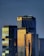 Several modern skyscrapers with reflective glass surfaces appear in the frame. The tallest building has the words 'The Ritz-Carlton' on it. The buildings are illuminated by what seems to be the golden light of either sunrise or sunset, giving them a warm glow against a darker background.