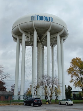 A tall, cylindrical water tower with the word 'Toronto' painted in blue at the top stands prominently against a cloudy sky. The structure is supported by multiple vertical pillars. In the foreground, there are a few leafless trees, two parked cars, and a brick building partially visible on the left side. The surrounding area is fenced, with some autumnal trees showing yellow and brown leaves.