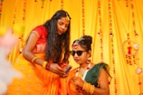 Two women in traditional Indian attire are in a festive setting decorated with bright yellow and colorful ornaments. One woman is holding a smartphone, while the other wears sunglasses and flower jewelry, suggesting a celebratory or wedding occasion.