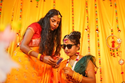 Two women in traditional Indian attire are in a festive setting decorated with bright yellow and colorful ornaments. One woman is holding a smartphone, while the other wears sunglasses and flower jewelry, suggesting a celebratory or wedding occasion.