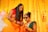 Two women in traditional Indian attire are in a festive setting decorated with bright yellow and colorful ornaments. One woman is holding a smartphone, while the other wears sunglasses and flower jewelry, suggesting a celebratory or wedding occasion.