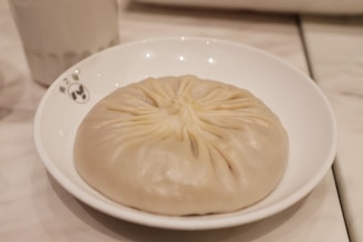 A large, round dumpling with neatly folded pleats on top sits on a white plate. The surface of the dumpling is smooth and slightly shiny, indicating it might be steamed. The background is softly blurred, showing a light-colored table setting.