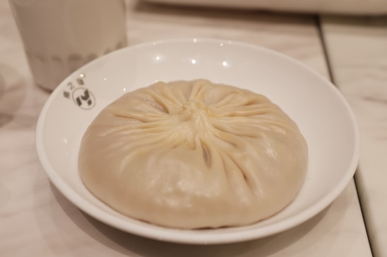 A large, round dumpling with neatly folded pleats on top sits on a white plate. The surface of the dumpling is smooth and slightly shiny, indicating it might be steamed. The background is softly blurred, showing a light-colored table setting.