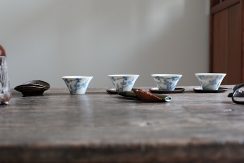Four small porcelain tea cups with blue floral designs are neatly placed in a row on a wooden table. Beside them lies a wooden tea scoop and other small utensils. The background is plain and minimalistic, creating a serene atmosphere.