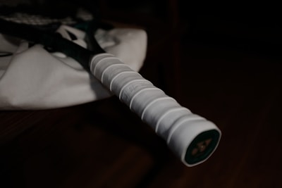 A close-up view of a tennis racket handle resting on what appears to be a white fabric. The focus is on the handle's grip, which is white and textured, contrasting against the dark background.