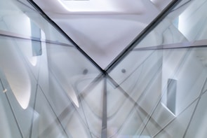 An abstract architectural structure featuring intersecting lines and reflective surfaces, creating a symmetrical pattern. The image incorporates translucent elements and soft gradients, contributing to a futuristic and minimalist design aesthetic.