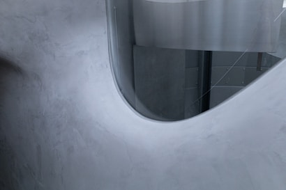 Curved, smooth concrete surfaces with a gray and metallic finish create a modern architectural interior. The design features a semi-transparent partition or window with rounded edges.
