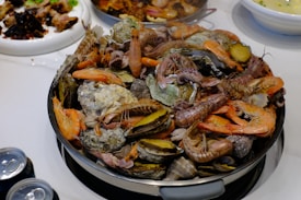 A large plate overflows with a variety of seafood, including shrimp, clams, oysters, and possibly langoustines, all arranged in an appetizing fashion. The dish is surrounded by other plates containing different food items, suggesting a lavish seafood feast.