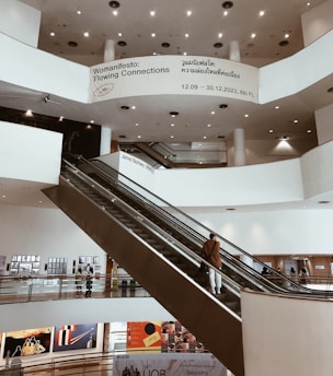 An indoor view of a multi-level art gallery with a modern design featuring large white walls, circular ceiling lights, and a prominent escalator. Several people are visible on the escalator and around the gallery, observing the artworks displayed on the walls. Banners and informational signs are present, including one advertising an exhibition called 'Womanifesto: Flowing Connections' with dates and floor information.