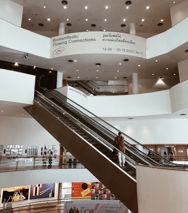 An indoor view of a multi-level art gallery with a modern design featuring large white walls, circular ceiling lights, and a prominent escalator. Several people are visible on the escalator and around the gallery, observing the artworks displayed on the walls. Banners and informational signs are present, including one advertising an exhibition called 'Womanifesto: Flowing Connections' with dates and floor information.