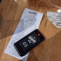 A receipt from Richiamo Coffee is placed on a wooden table. The receipt includes several items such as an ice caffe latte and various deserts. Beside the receipt is a wireless pager with the number 03 displayed and a small red light indicating it's active.