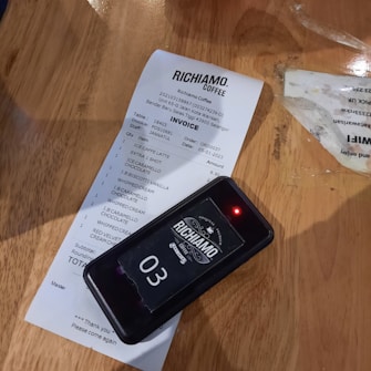 A receipt from Richiamo Coffee is placed on a wooden table. The receipt includes several items such as an ice caffe latte and various deserts. Beside the receipt is a wireless pager with the number 03 displayed and a small red light indicating it's active.