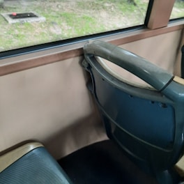 Interior of a vehicle featuring a row of blue and beige bus seats next to a window. Outside, there is a grassy area with a small concrete structure visible through the window.