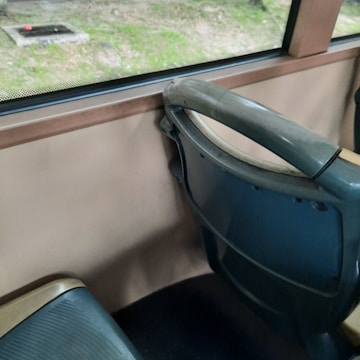 Interior of a vehicle featuring a row of blue and beige bus seats next to a window. Outside, there is a grassy area with a small concrete structure visible through the window.