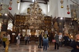 A richly decorated interior of a church featuring numerous ornate chandeliers hanging from the ceiling. The altar area is adorned with intricate woodwork and religious icons. Several people are standing and walking around, some taking photos or admiring the surroundings. The walls are embellished with stained glass and religious paintings.