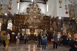 A richly decorated interior of a church featuring numerous ornate chandeliers hanging from the ceiling. The altar area is adorned with intricate woodwork and religious icons. Several people are standing and walking around, some taking photos or admiring the surroundings. The walls are embellished with stained glass and religious paintings.
