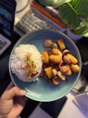 A plate of food featuring a mound of white rice garnished with fried shallots and chopped green and red chilies on one side. The other side has an assortment of fried items including sausages, crispy golden-brown nuggets, and vegetables. A large green leaf is visible in the top right corner, casting a shadow over part of the plate. The background shows a blurred laptop and other items on a workspace.