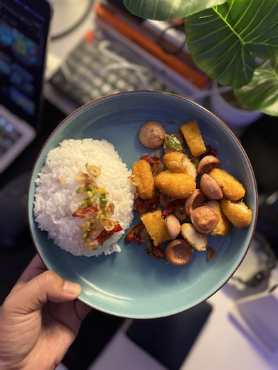 A plate of food featuring a mound of white rice garnished with fried shallots and chopped green and red chilies on one side. The other side has an assortment of fried items including sausages, crispy golden-brown nuggets, and vegetables. A large green leaf is visible in the top right corner, casting a shadow over part of the plate. The background shows a blurred laptop and other items on a workspace.