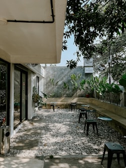 An outdoor seating area with a gravel floor, several small black stools, and circular tables. The area is flanked by a low wall with potted plants. A building with large glass windows is on the left, and overhanging foliage provides some shade. Signs and trees are visible in the background.