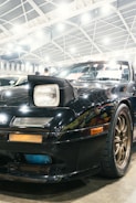 A close-up view of a black sports car with distinctly visible headlights. The car is parked indoors, under a large, brightly lit roof structure with metal beams. Reflections and details on the car's surface indicate it is well-polished. Bronze-colored wheels are partially visible, adding a contrast to the car’s dark body.
