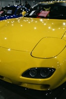 A brightly colored yellow car with a sleek and polished finish, parked indoors. The hood features striking headlights and the windshield displays a sign in multiple languages cautioning not to touch the vehicle. Reflections of lights on the car's surface add to the shiny aesthetic.